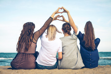 four girls sit on the seashore and show their hands a heart sign over their heads
