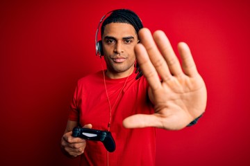 African american man with dreadlocks playing video game using joystick and headphones with open hand doing stop sign with serious and confident expression, defense gesture