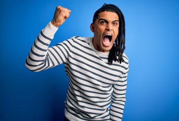 Young handsome african american afro man with dreadlocks wearing casual striped sweater angry and mad raising fist frustrated and furious while shouting with anger. Rage and aggressive concept.