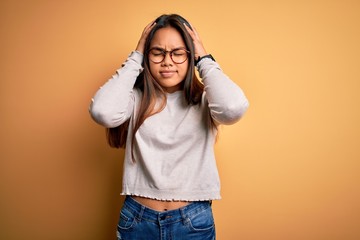 Young beautiful asian girl wearing casual sweater and glasses over yellow background suffering from headache desperate and stressed because pain and migraine. Hands on head.