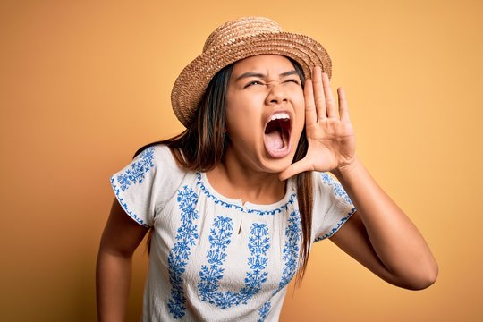 Young beautiful asian girl wearing casual t-shirt and hat standing over yellow background shouting and screaming loud to side with hand on mouth. Communication concept.