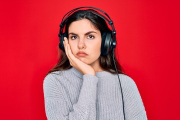 Young beautiful girl wearing modern headphones listening to music over red background thinking looking tired and bored with depression problems with crossed arms.