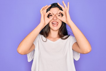 Obraz na płótnie Canvas Young beautiful brunette woman wearing casual white t-shirt over purple background doing ok gesture like binoculars sticking tongue out, eyes looking through fingers. Crazy expression.