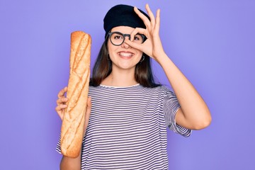 Young beautiful girl wearing fashion french beret holding fresh baked bread baguette with happy face smiling doing ok sign with hand on eye looking through fingers