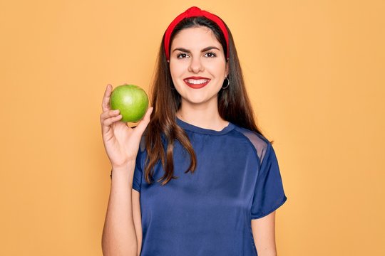 Young beautiful girl eating fresh organic healthy green apple over yellow background with a happy face standing and smiling with a confident smile showing teeth