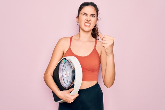 Young beautiful fitness woman wearing sport clothes holding weighting machine scale annoyed and frustrated shouting with anger, crazy and yelling with raised hand, anger concept