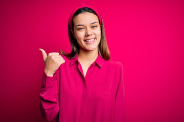 Young beautiful brunette girl wearing casual shirt standing over isolated pink background smiling with happy face looking and pointing to the side with thumb up.