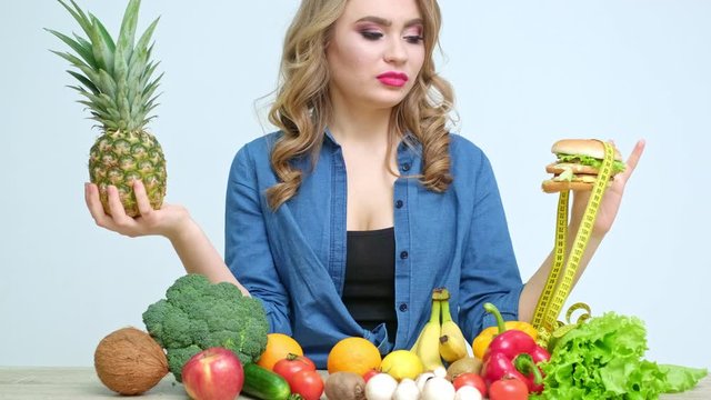 Pretty woman in the kitchen holding a malicious burger on a background of fresh vegetables and fruits