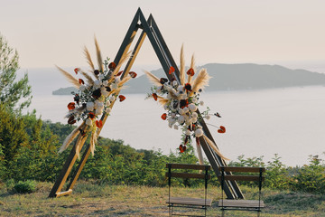 Original wedding arch on top of the mountain with white and red flowers at sunset. Romantic ceremony in the style of boho