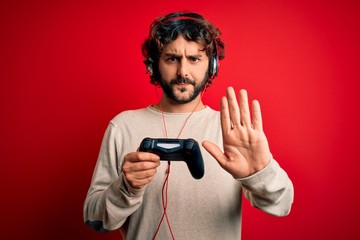 Young gamer man with curly hair and beard playing video game using joystick and headphones with open hand doing stop sign with serious and confident expression, defense gesture