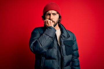 Young handsome man with beard wearing casual coat and wool cap over red background looking stressed and nervous with hands on mouth biting nails. Anxiety problem.