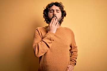 Young handsome man with beard wearing casual sweater standing over yellow background bored yawning tired covering mouth with hand. Restless and sleepiness.