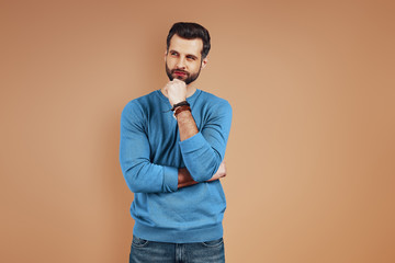 Thoughtful young man in casual wear looking away and keeping hand on chin while standing against brown background