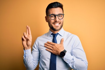 Young handsome businessman wearing tie and glasses standing over yellow background smiling swearing with hand on chest and fingers up, making a loyalty promise oath