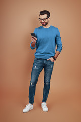 Full length of happy young man in smart casual wear using smart phone and smiling while standing against brown background