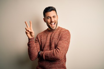 Young handsome man wearing casual sweater standing over isolated white background smiling with happy face winking at the camera doing victory sign with fingers. Number two.