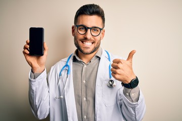 Young doctor man wearing stethoscope showing smartphone screen over isolated background happy with big smile doing ok sign, thumb up with fingers, excellent sign