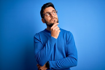 Young handsome man with beard wearing casual sweater and glasses over blue background with hand on chin thinking about question, pensive expression. Smiling with thoughtful face. Doubt concept.