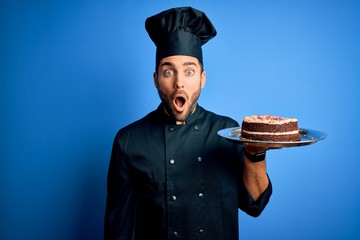 Young handsome cooker man with beard wearing uniform and hat holding tray with cake scared in shock...