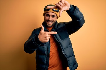 Young handsome skier man with beard wearing snow sportswear and ski goggles smiling making frame with hands and fingers with happy face. Creativity and photography concept.