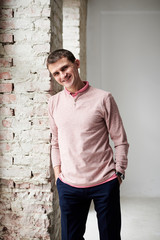 Obraz premium Young brunette man, wearing casual light pink jumper, leaning on white brick wall in light room, smiling, laughing.Happy emotions. Three-quarter portrait of businessman, resting on his day off.