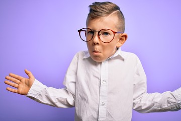 Young little caucasian kid with blue eyes wearing glasses and white shirt over purple background clueless and confused expression with arms and hands raised. Doubt concept.