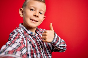 Close up of young little caucasian kid with blue eyes taking a selfie photo over red background happy with big smile doing ok sign, thumb up with fingers, excellent sign