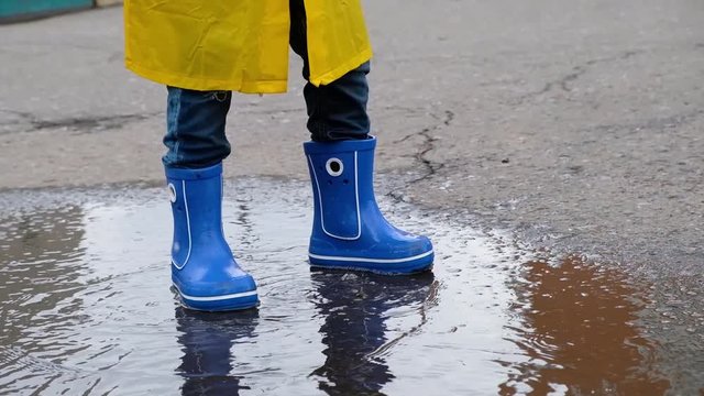 Funny child kid in a yellow raincoat and rubber boots jumping in a puddle, close up