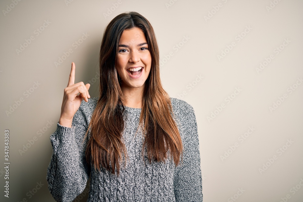 Poster Young beautiful girl wearing casual sweater standing over isolated white background pointing finger up with successful idea. Exited and happy. Number one.