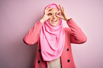 Young beautiful girl wearing muslim hijab standing over isolated pink background doing ok gesture like binoculars sticking tongue out, eyes looking through fingers. Crazy expression.