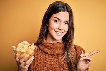 Young beautiful girl holding bowl with chips potatoes standing over yellow background very happy pointing with hand and finger to the side