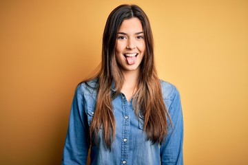 Young beautiful girl wearing casual denim shirt standing over isolated yellow background sticking tongue out happy with funny expression. Emotion concept.