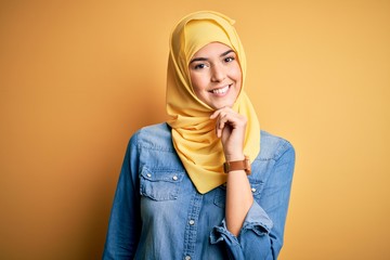 Young beautiful girl wearing muslim hijab standing over isolated yellow background looking confident at the camera with smile with crossed arms and hand raised on chin. Thinking positive.