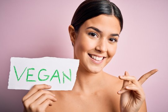Young beautiful girl holding paper with vegan message over isolated pink background very happy pointing with hand and finger to the side
