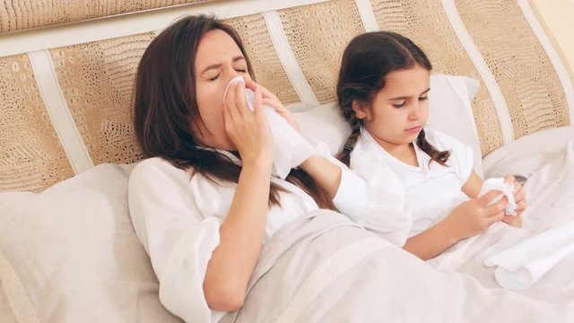 Mom and daughter lie together in bed and get sick, blow their nose, sneeze. The family is sick, sitting at home