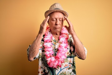 Grey haired senior man wearing summer hat and hawaiian lei over yellow background with hand on head...