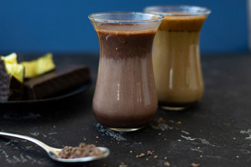 Dalgona coffee and cocoa in glass transparent glasses. Nearby lies a metal spoon with instant coffee, and a chocolate dessert on a saucer