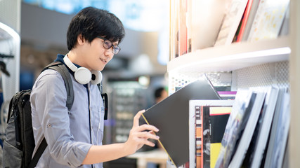 Fototapeta premium Asian man student choosing and picking book from modern bookshelf in bookstore. Buying bestseller textbook collection at book shop. Education and learning concept