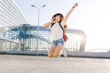 Naklejka premium Funny photo of beautiful excited young asian woman in jeans shorts and white t-shirt, with red backpack, talking phone, and jumping up with one hand raised, waiting for journey near modern airport