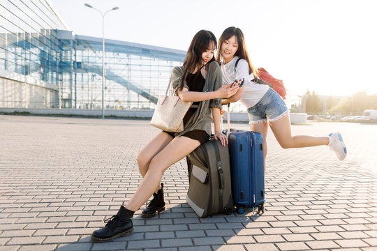 Young pretty satisfied asian girls showing photos or flight information on smartphone to her pretty best friend, while sitting outdoors on suitcase on the airport terminal background
