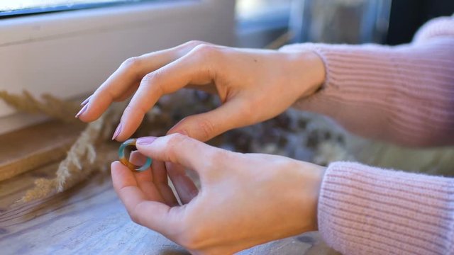 Female hands with natural manicure are showing orange and green gemstone rings made from agate and opal gems on wooden windowsill with spikelets on it