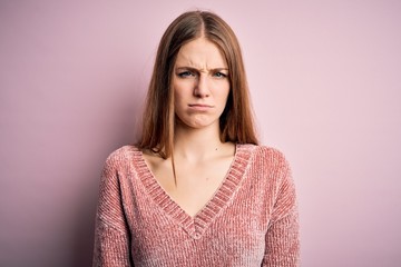Young beautiful redhead woman wearing casual sweater over isolated pink background skeptic and...