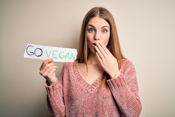 Young beautiful redhead woman asking for veggie food holding paper with vegan message cover mouth...