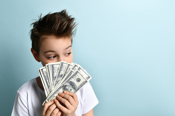 Stylish boy keeping fan of cash dollars in hand wondering how to invest or spend them better. Half-length portrait isolated on light blue