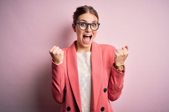 Young beautiful redhead woman wearing jacket and glasses over isolated pink background celebrating surprised and amazed for success with arms raised and open eyes. Winner concept.
