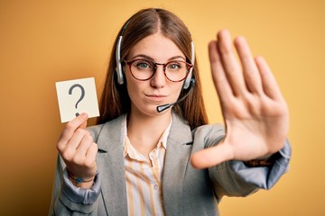 Young redhead call center agent woman using headset holding question mark reminder with open hand doing stop sign with serious and confident expression, defense gesture