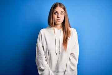 Young beautiful redhead sporty woman wearing sweatshirt over isolated blue background puffing cheeks with funny face. Mouth inflated with air, crazy expression.