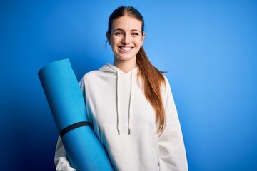Young beautiful redhead sporty woman doing sport holding yoga mat over blue background with a happy face standing and smiling with a confident smile showing teeth