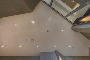 look up on suspended ceiling with halogen spots lamps and drywall construction with fire alarm sensor in empty room in apartment or house. Stretch ceiling white and complex shape.