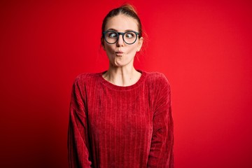 Young beautiful redhead woman wearing casual sweater over isolated red background making fish face with lips, crazy and comical gesture. Funny expression.
